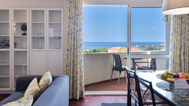 Ein Zimmer mit einer Couch, einem Esstisch mit Stühlen, einem Glasvitrinen und Blick aufs Meer.