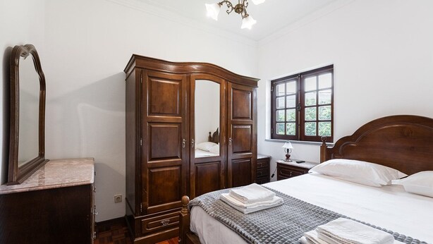 Un dormitorio con un armario de madera, una cama con ropa de cama blanca, una mesita de noche y una ventana con vistas a zonas verdes.