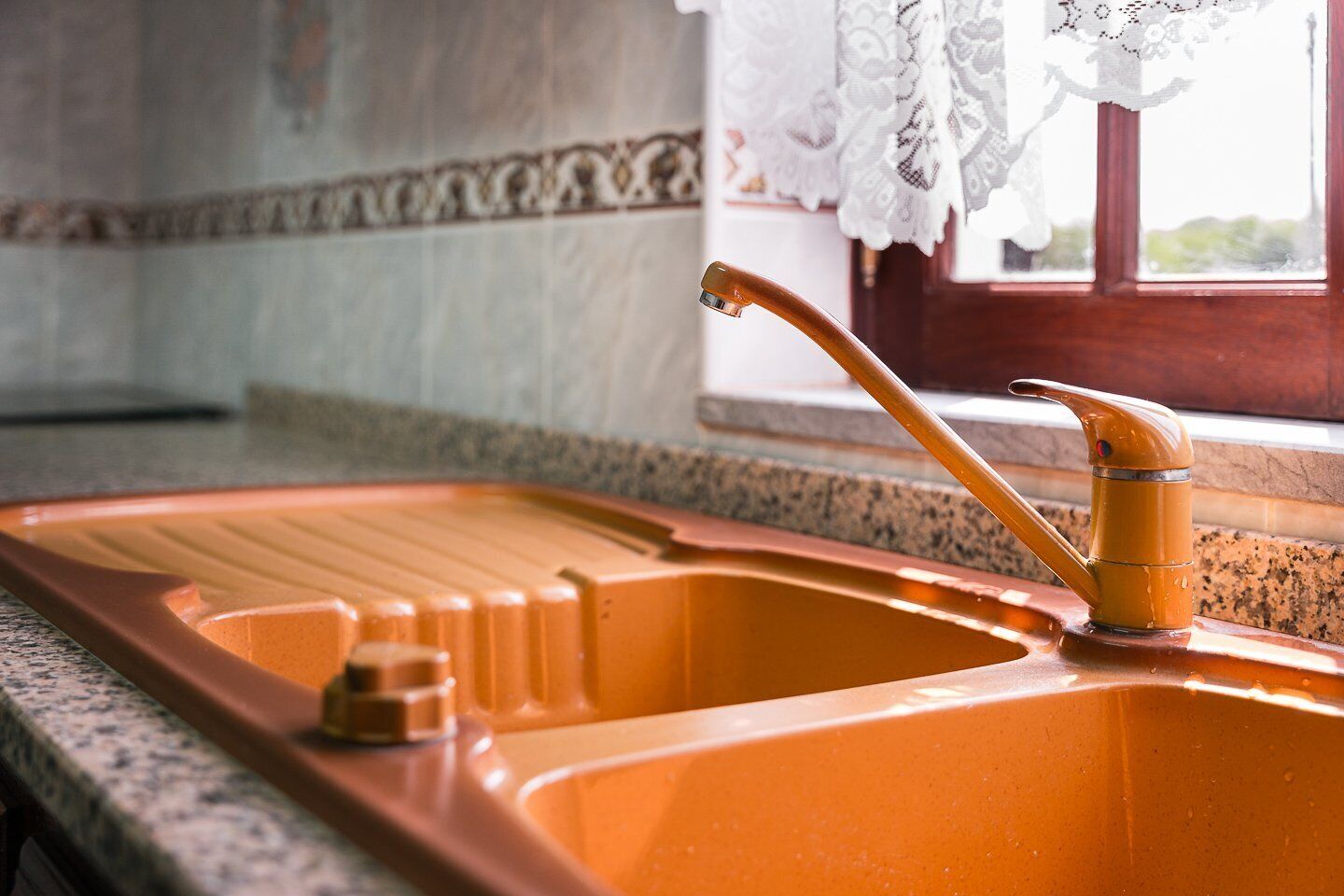 A kitchen sink with a single faucet, a soap dish, and a window with lace curtains.