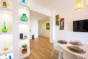 Ein moderner Essbereich mit einem weißen Tisch für zwei Personen, einem an der Wand befestigten Flachbildfernseher und einem Flur, der in ein weiteres Zimmer führt.