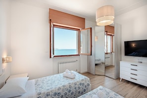 A bedroom with two beds, a large mirror, a TV, and a view of the sea.