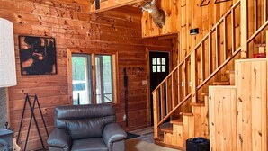 A cozy log cabin interior with a leather sofa, a round wooden coffee table, and a staircase leading to a loft area. - Log Cabin w/ Hot Tub - close to Whiteface (Wilmington)