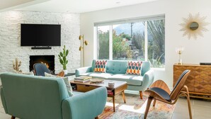 Uma sala de estar moderna com lareira de tijolos brancos, uma televisão de tela plana montada acima, um sofá azul claro com almofadas coloridas, uma mesa de centro de madeira e uma grande janela com vista para o verde lá fora.