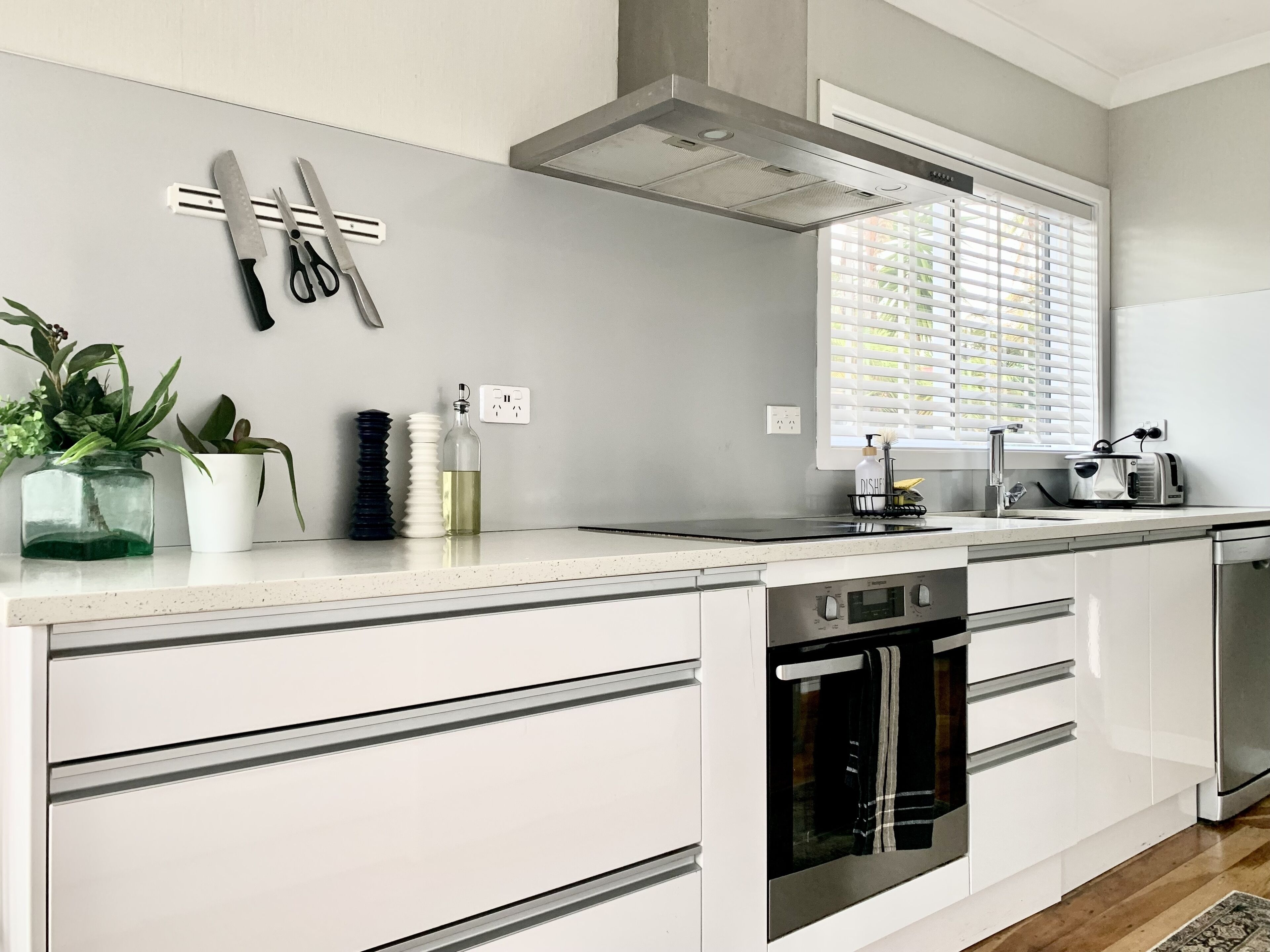 Una cocina moderna con armarios blancos, horno empotrado, lavavajillas y una ventana con persianas.