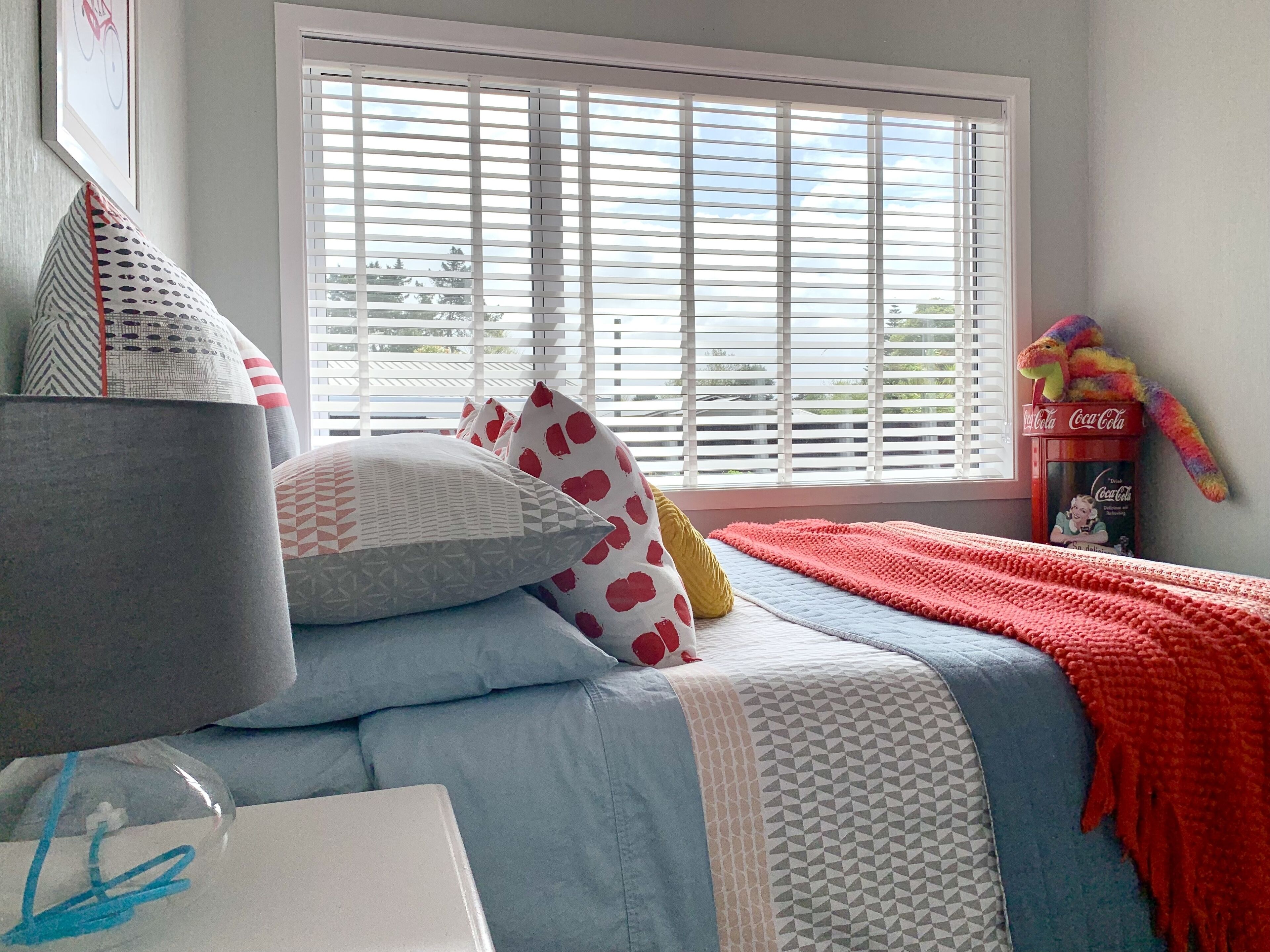 A bedroom with a bed, pillows, a blanket, a window with blinds, and a stuffed toy.