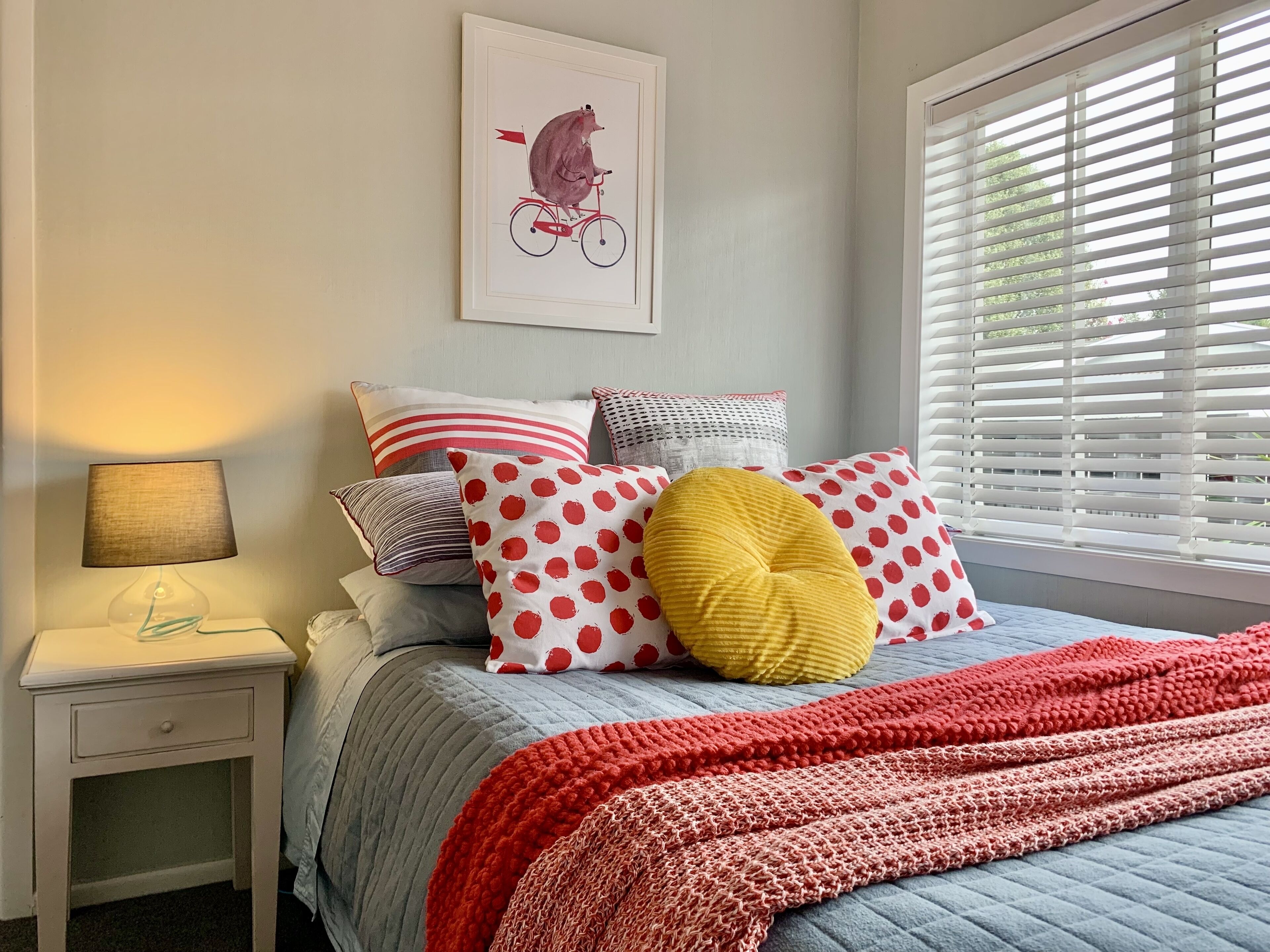 A bedroom with a bed, pillows, a bedside table, a lamp, and a window with blinds.