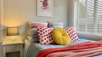 A bedroom with a bed, pillows, a bedside table, a lamp, and a window with blinds.