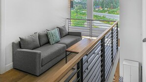 A modern living room with a grey sofa, a wooden bench, and large windows offering a view of greenery and mountains. - New! Modern Ski-in/Ski-out Mountain View Chalet by Village + Hot Tub/EV Charging (Snoqualmie Pass)