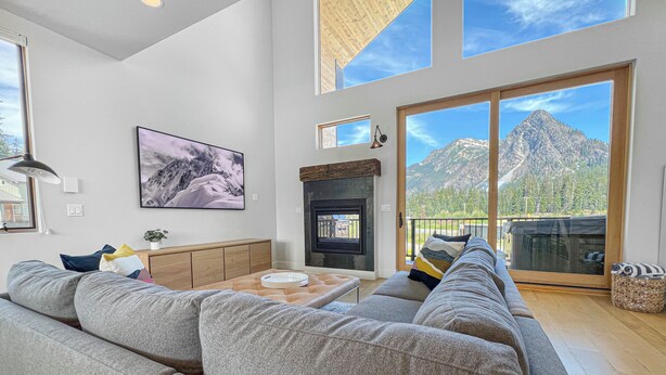 A spacious living room with a large gray sectional sofa, a fireplace, and a mountain view through large sliding glass doors. - New! Modern Ski-in/Ski-out Mountain View Chalet by Village + Hot Tub/EV Charging (Snoqualmie Pass)