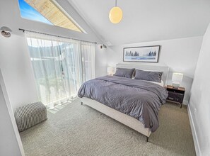 Et soveværelse med en stor seng, ovenlys, et indrammet billede på væggen og et sengebord med en lampe.
