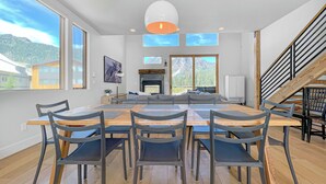 A modern dining area with a large wooden table and chairs, a fireplace, and a view of mountains through large windows. - New! Modern Ski-in/Ski-out Mountain View Chalet by Village + Hot Tub/EV Charging (Snoqualmie Pass)