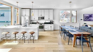 A modern kitchen with white cabinets, a large island with stools, a dining area with a long table and chairs, and a view of a house through the window. - New! Modern Ski-in/Ski-out Mountain View Chalet by Village + Hot Tub/EV Charging (Snoqualmie Pass)