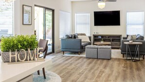A modern living room with a flat-screen TV, grey sofas, a coffee table, and a potted plant with the word "HOME" on it. - Resort Backyard with Pool, Spa & Putting Green (Chandler)