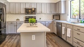 A modern kitchen with grey cabinets, a central island, stainless steel appliances, and a window providing natural light. - Resort Backyard with Pool, Spa & Putting Green (Chandler)