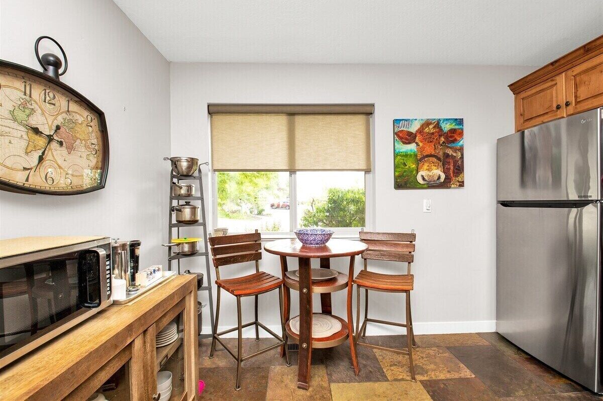 Une cuisine avec une table en bois, des chaises, une horloge, un réfrigérateur et une fenêtre donnant sur un paysage verdoyant.