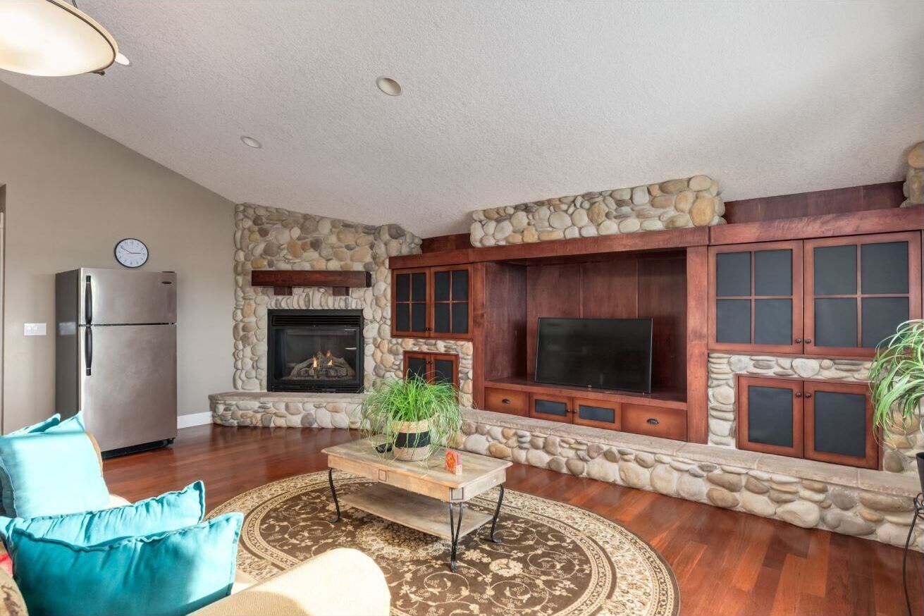 A living room with a stone fireplace, wooden cabinets, a flat-screen TV, and a refrigerator.