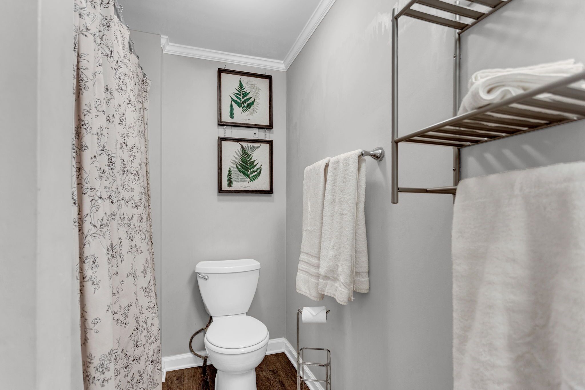 A bathroom with a toilet, a shower curtain, a towel rack with towels, and two framed botanical prints on the wall.
