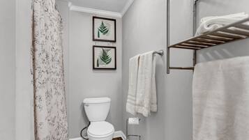 A bathroom with a toilet, a shower curtain, a towel rack with towels, and two framed botanical prints on the wall.