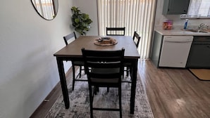 A dining area with a wooden table, chairs, a rug, a potted plant, and a kitchen in the background. - COZY 2Bedroom/1Bath Duplex  (Enid)