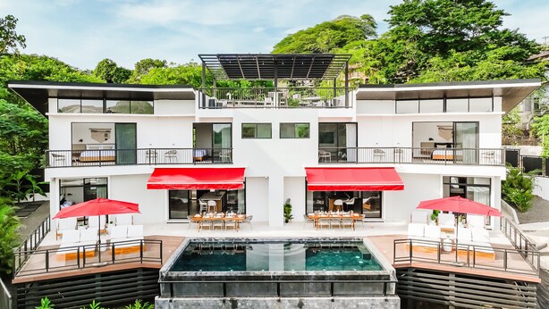 A modern two-story building with a rooftop terrace, a swimming pool, and outdoor seating areas with red umbrellas. - Elegant Modern Villa with Private Pool & Rooftop, Near Beach and City Center (Tamarindo)