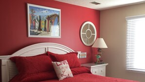 A bedroom with a red bedspread, a white headboard, a framed picture on the wall, a bedside table with a lamp, and a window with blinds. - Rhue's Haven less than 5 minutes from the most wonderful beach in NC (Sunset Beach)