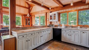 A spacious kitchen with granite countertops, white cabinetry, and a large island. There are multiple windows providing natural light and views of the surrounding forest.