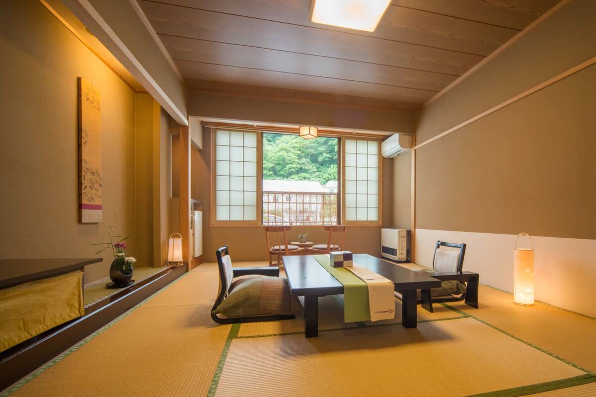 A traditional Japanese room with tatami flooring, a low table, chairs, a TV, and a window with a view of greenery.