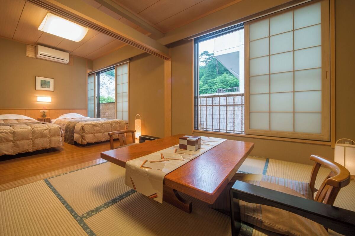 A traditional Japanese room with two beds, a wooden table, tatami mats, and large windows with a view of greenery.