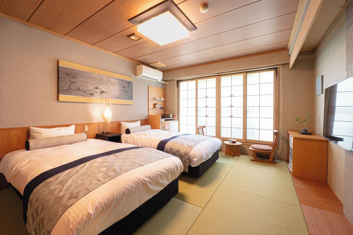 A hotel room with two beds, a painting, a window with a view, and a small table.