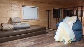 Una habitación acogedora con piso de madera, un sofá con cojines, un sillón de peluche y una ventana con persianas. Se observa una escalera con barandilla.