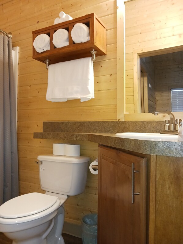 Un baño de madera con inodoro, lavamanos y espejo. Toallas cuelgan de un estante sobre el inodoro.