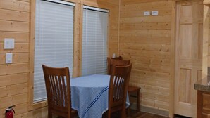 Interior de una cabaña de madera con una mesa de comedor y sillas, dos ventanas con persianas, techo de madera y un extintor en el suelo.