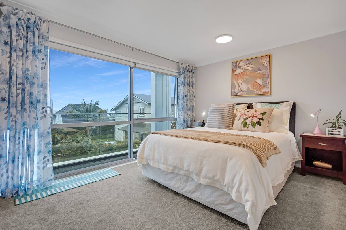 A bedroom with a large window, a bed with a beige blanket, a nightstand with a lamp, and a framed artwork on the wall.