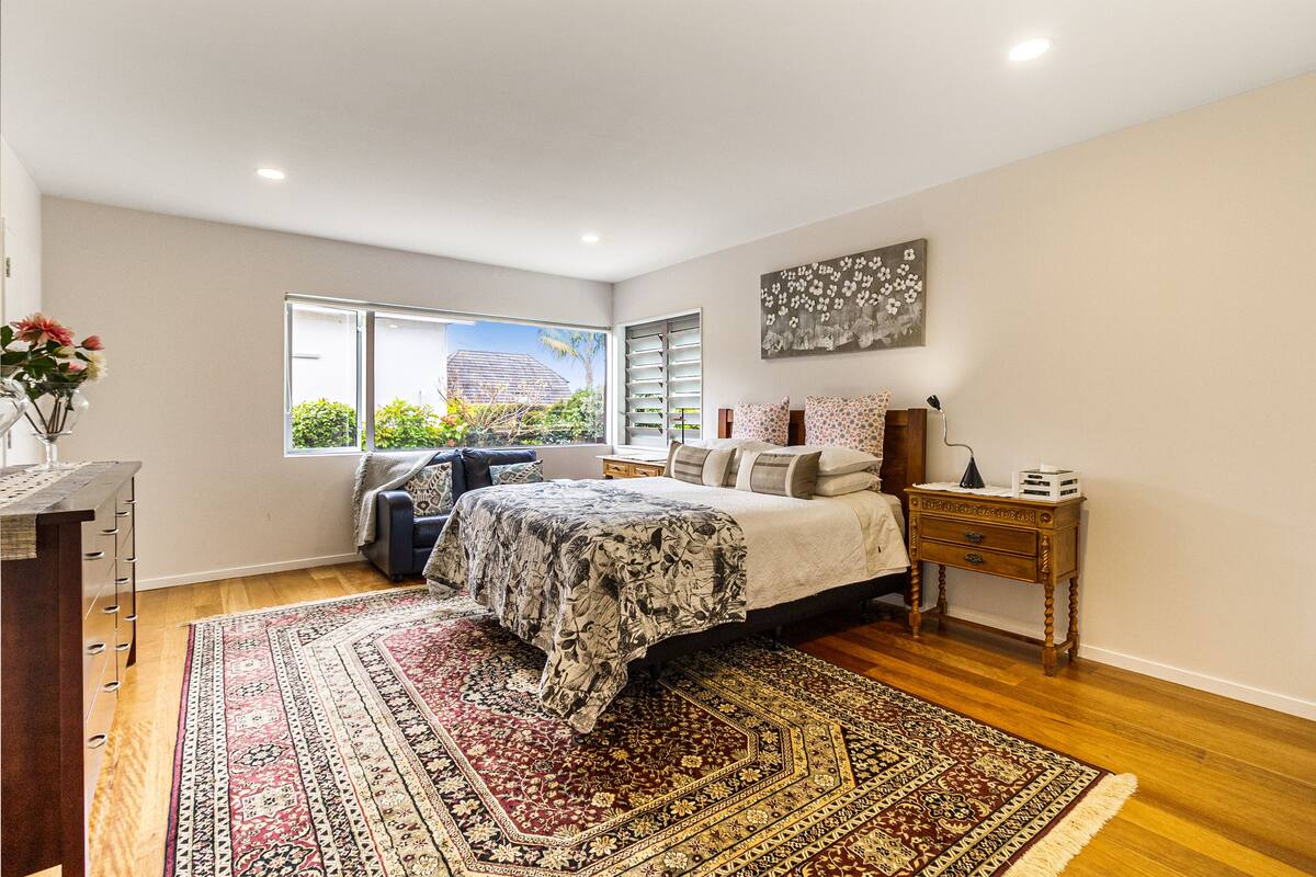 A bedroom with a large bed, a nightstand, a dresser, a window with a view of greenery, and a decorative wall piece.
