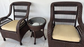 Dos sillas de mimbre con cojines color beige, una mesa de cristal y una puerta blanca con un ventanal arriba.