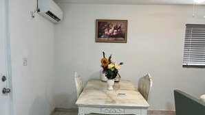 Un comedor con una mesa de mármol, sillas, un jarrón de flores, un cuadro y una ventana con persianas.