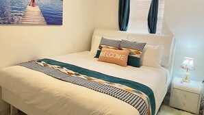Un dormitorio con cama, una mesita de noche, una lámpara, un cuadro y una ventana con cortinas.