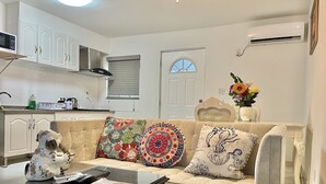 Una sala de estar con un sofá, una mesa de centro y un perro de cerámica decorativo. Hay una ventana con persianas, una puerta y un equipo de aire acondicionado en la pared.