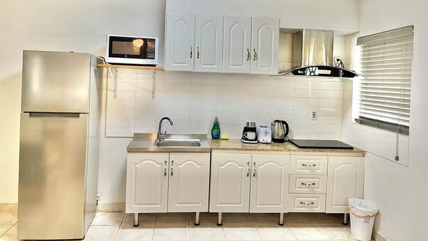 Una cocina compacta con armarios blancos, un refrigerador de acero inoxidable, un microondas, un fregadero y una ventana con persianas.