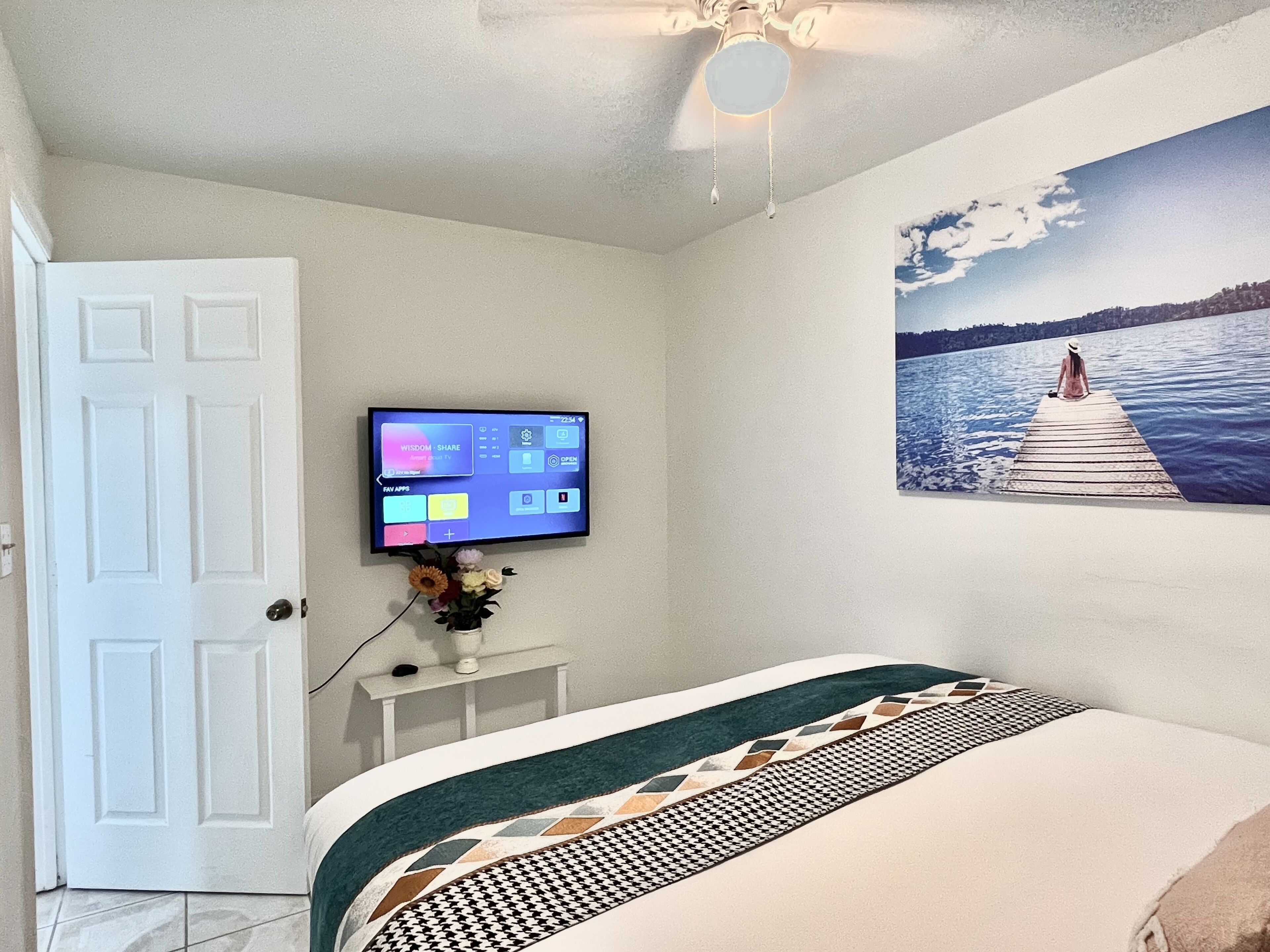 A bedroom with a bed, a television on the wall, a painting of a lake, and a ceiling fan.