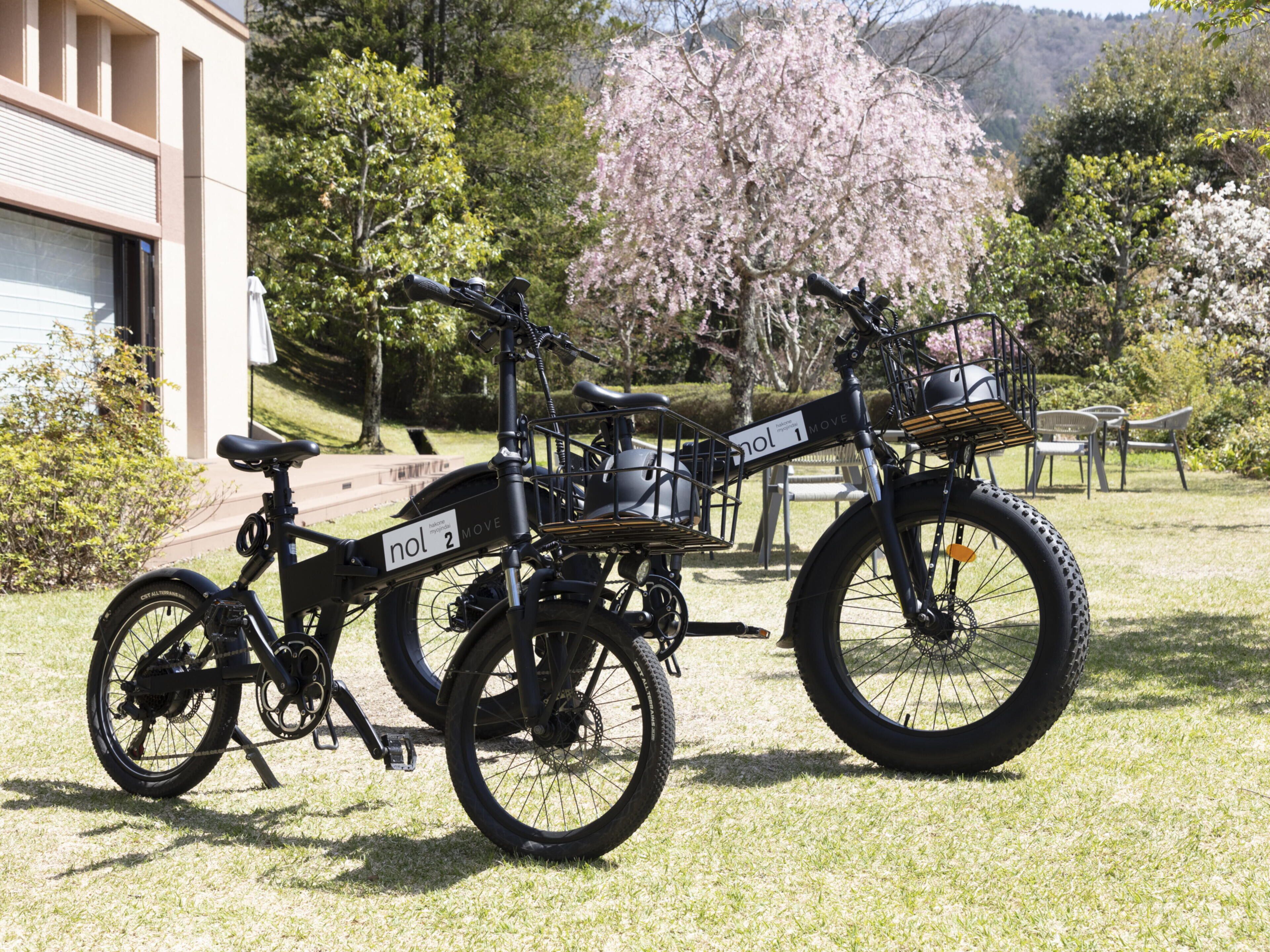 Two stationary bicycles with 'nol 2 MOVE' branding, a building with a closed shutter, and cherry blossom trees in the background.