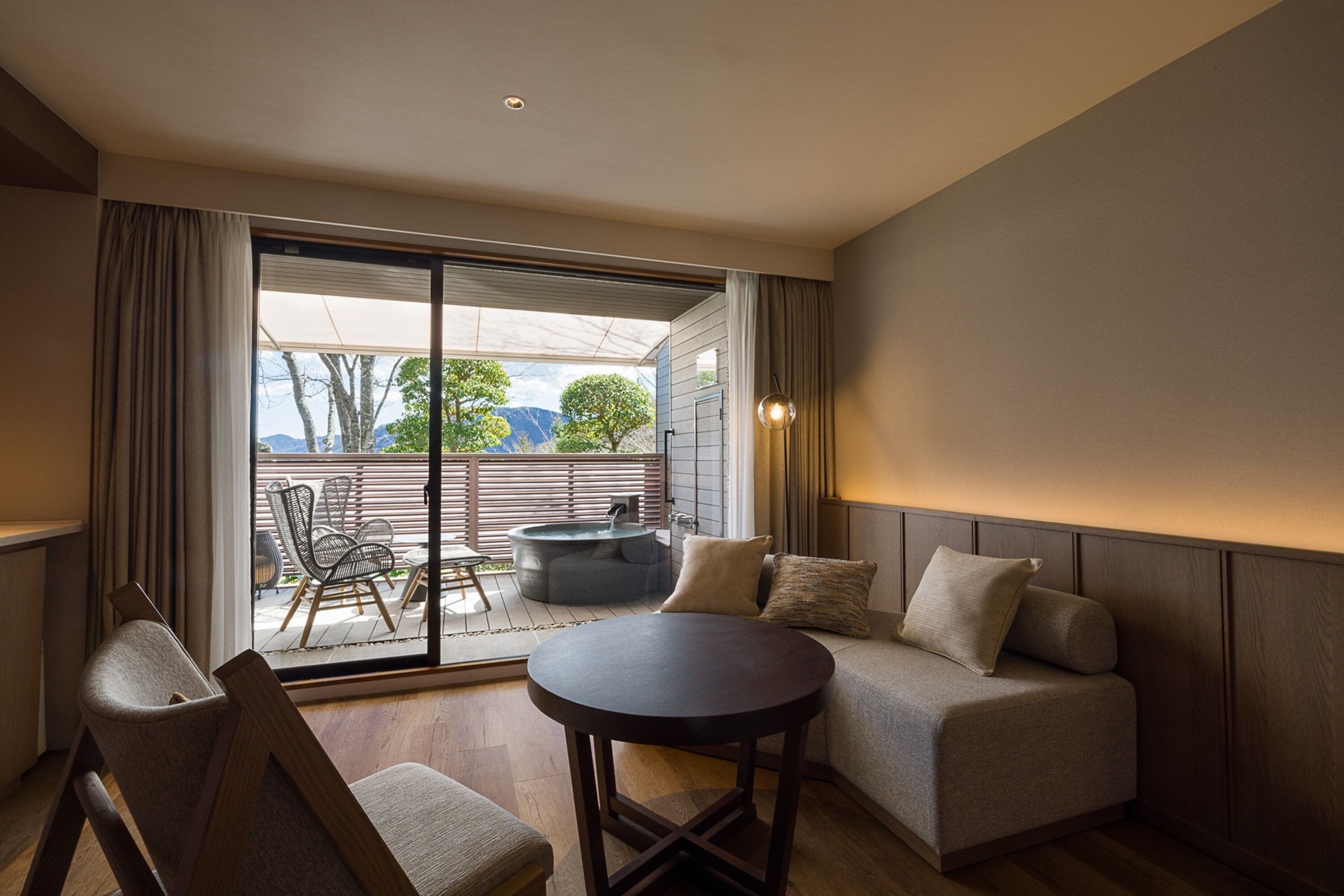 A modern living room with a sofa, a round table, and a sliding glass door leading to an outdoor seating area with a hot tub.