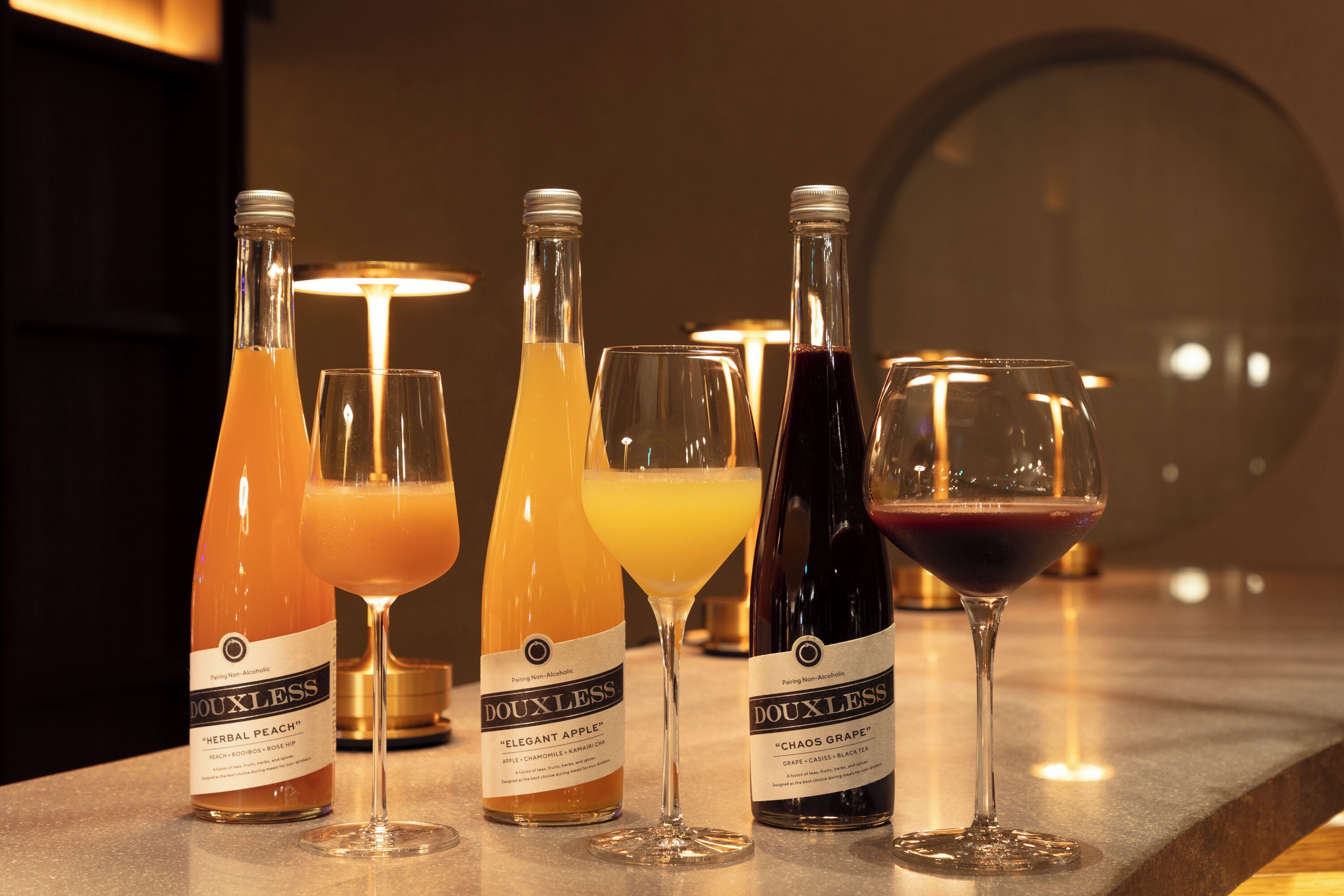 A bar setting with three bottles of Douxless beverages and three glasses, each containing a different drink.