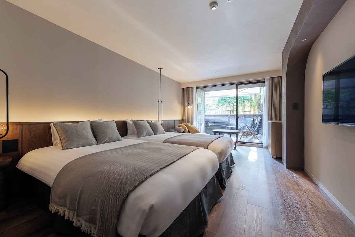 Quarto de hotel moderno com duas camas grandes, uma varanda com mesa e cadeiras, e uma janela ampla.