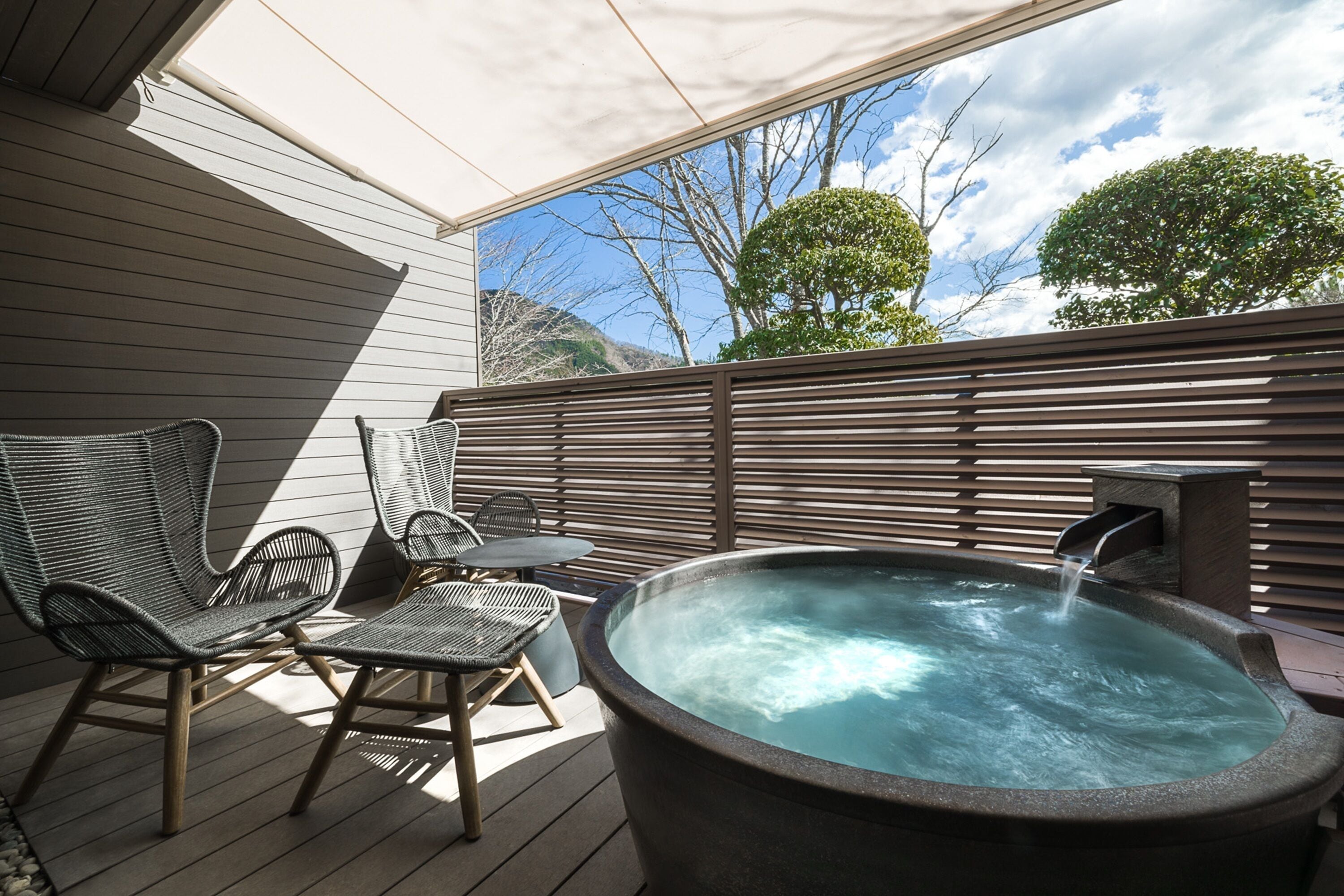 A hot tub on a wooden deck with wicker chairs and a table, overlooking a mountainous landscape.