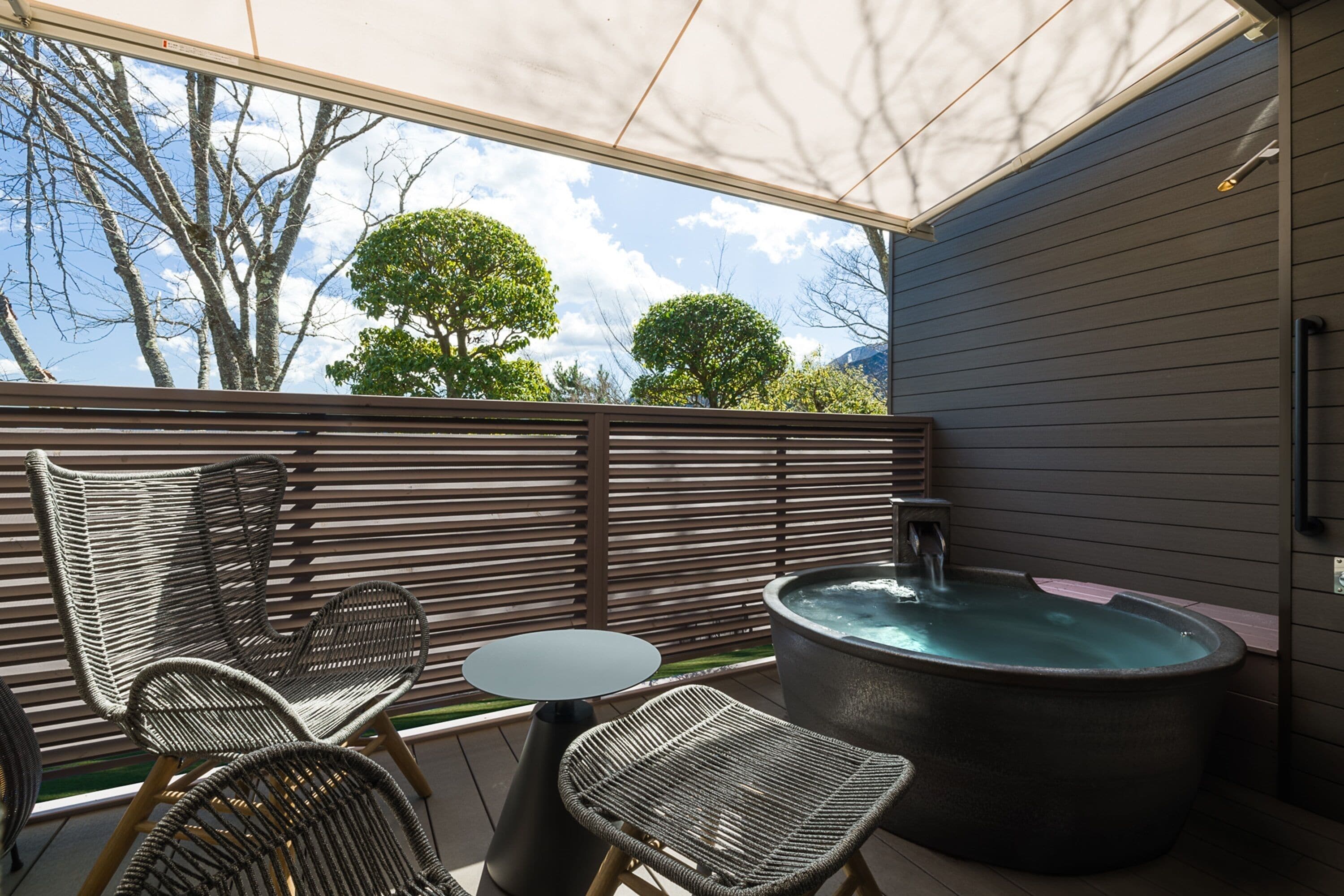 An outdoor seating area with wicker chairs and a table, a hot tub, and a view of trees and sky.