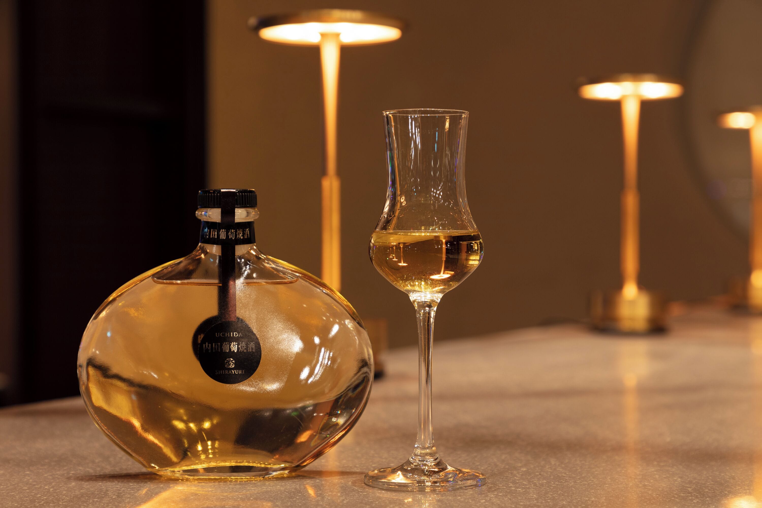 Eine Flasche UCHIDA Shirayuri Sake und ein Sake-Glas auf einem Tisch, im Hintergrund eine warme Lichtquelle.