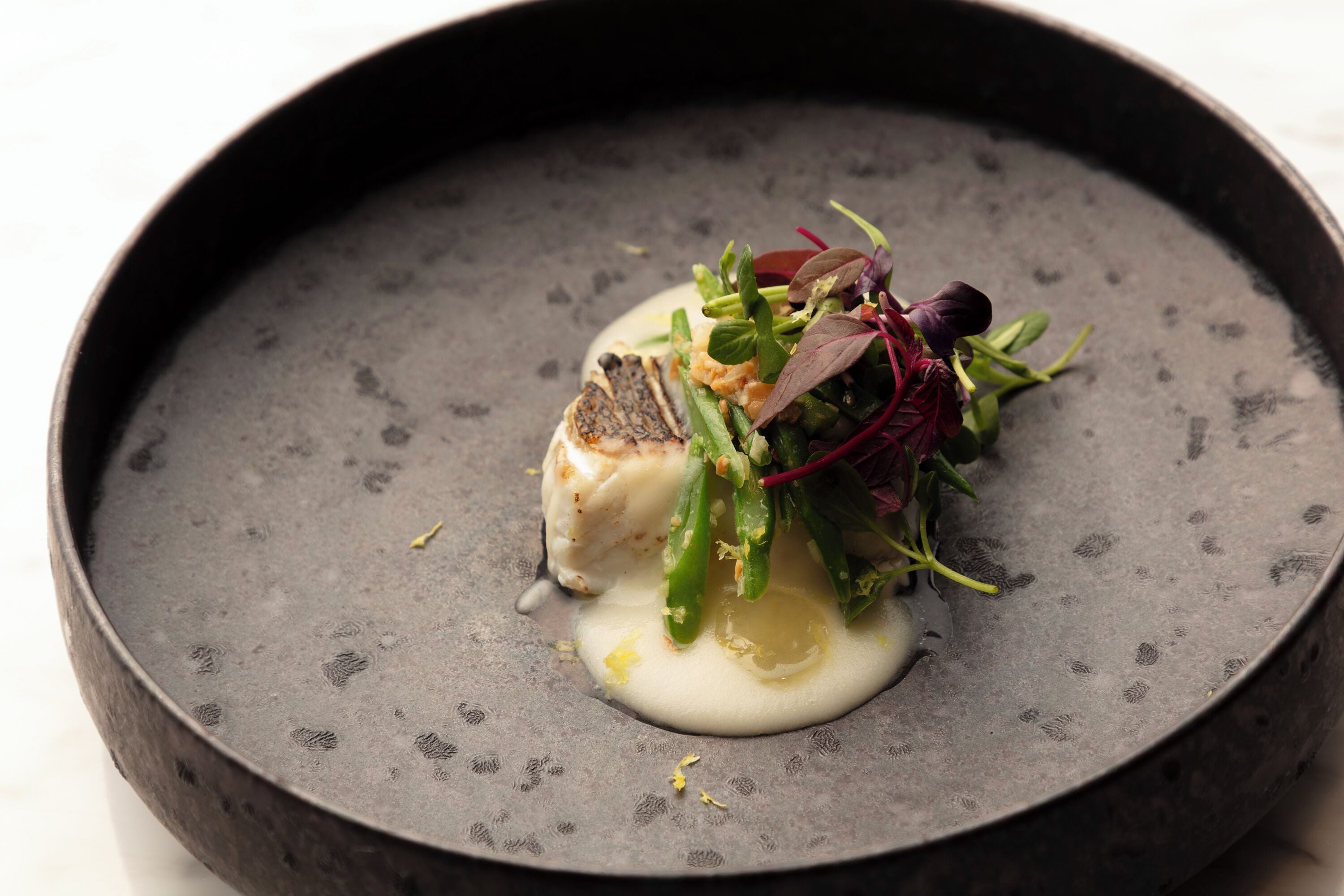 Un plat composé de poisson, de légumes verts et d’une sauce crémeuse, le tout servi dans une assiette foncée.