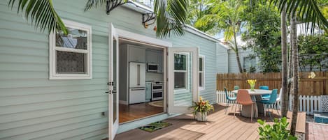 Una casa azul claro con terraza de madera, una pequeña cocina y un comedor con mobiliario exterior.