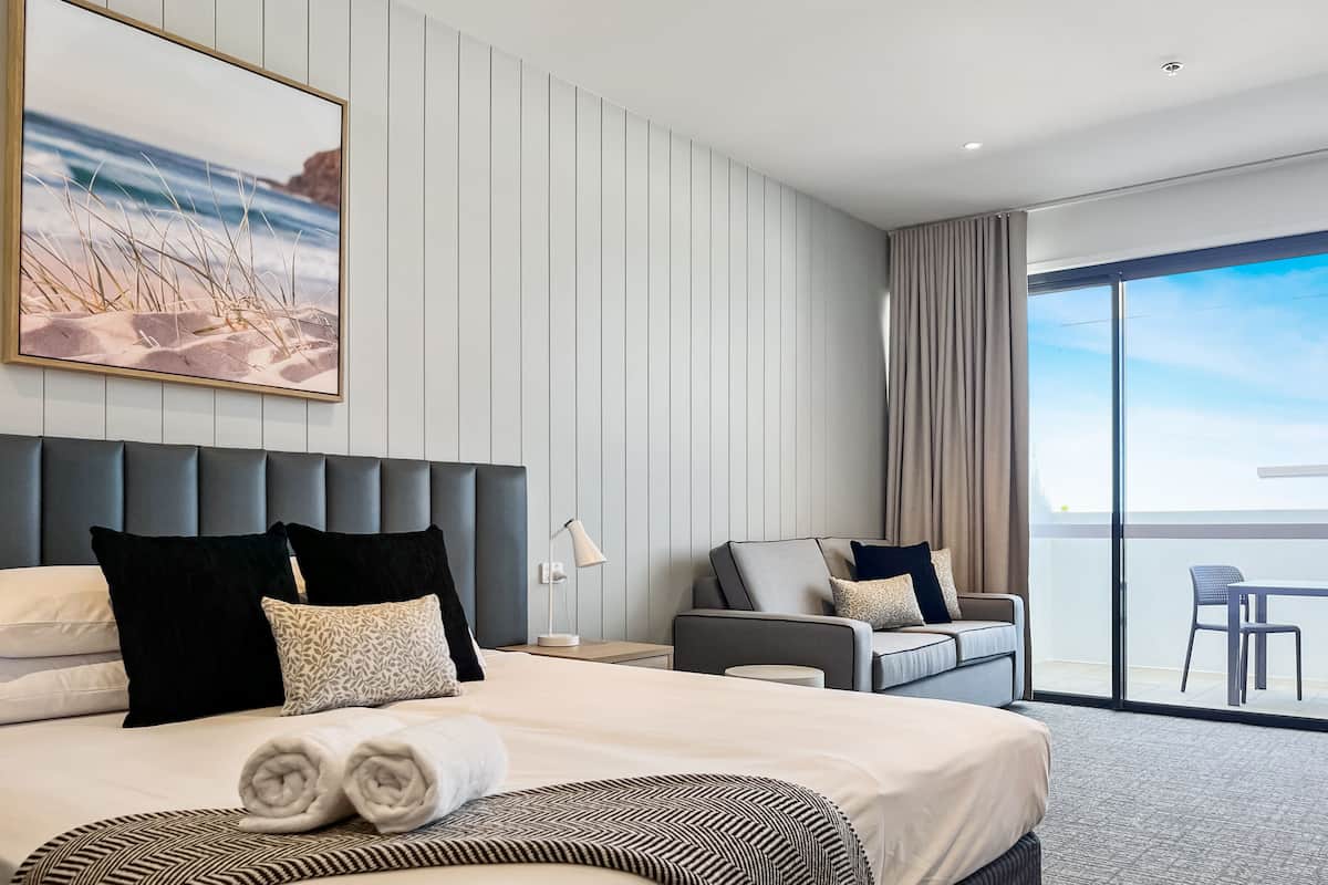 Una habitación de hotel moderna con una cama grande, un sofá, una zona de comedor y un balcón con vistas.